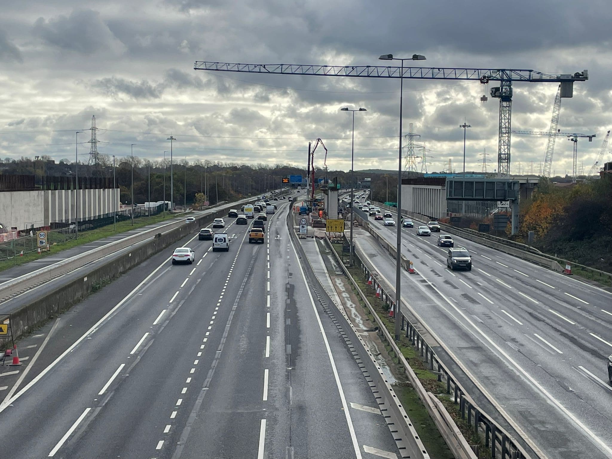 A live UK motorway scheme under an overcast sky — narrow lanes, cones, a concrete pump working the central reserve, tower cranes behind